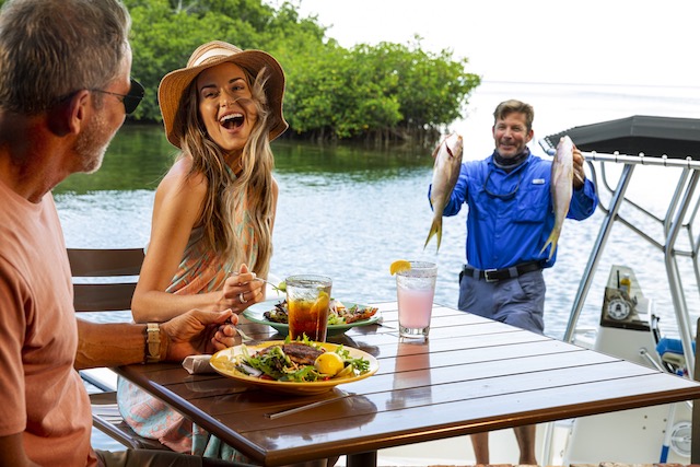 A couple dining dockside while a man in the background holds up two freshly-caught fish