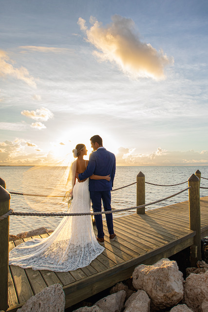 A bride and groom standing on a pier at sunset