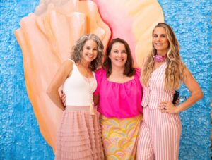 Nadine Grossman Orr, Liz Love and Kirby Myers posing in front a mural with a blue background and pink & orange conch shell